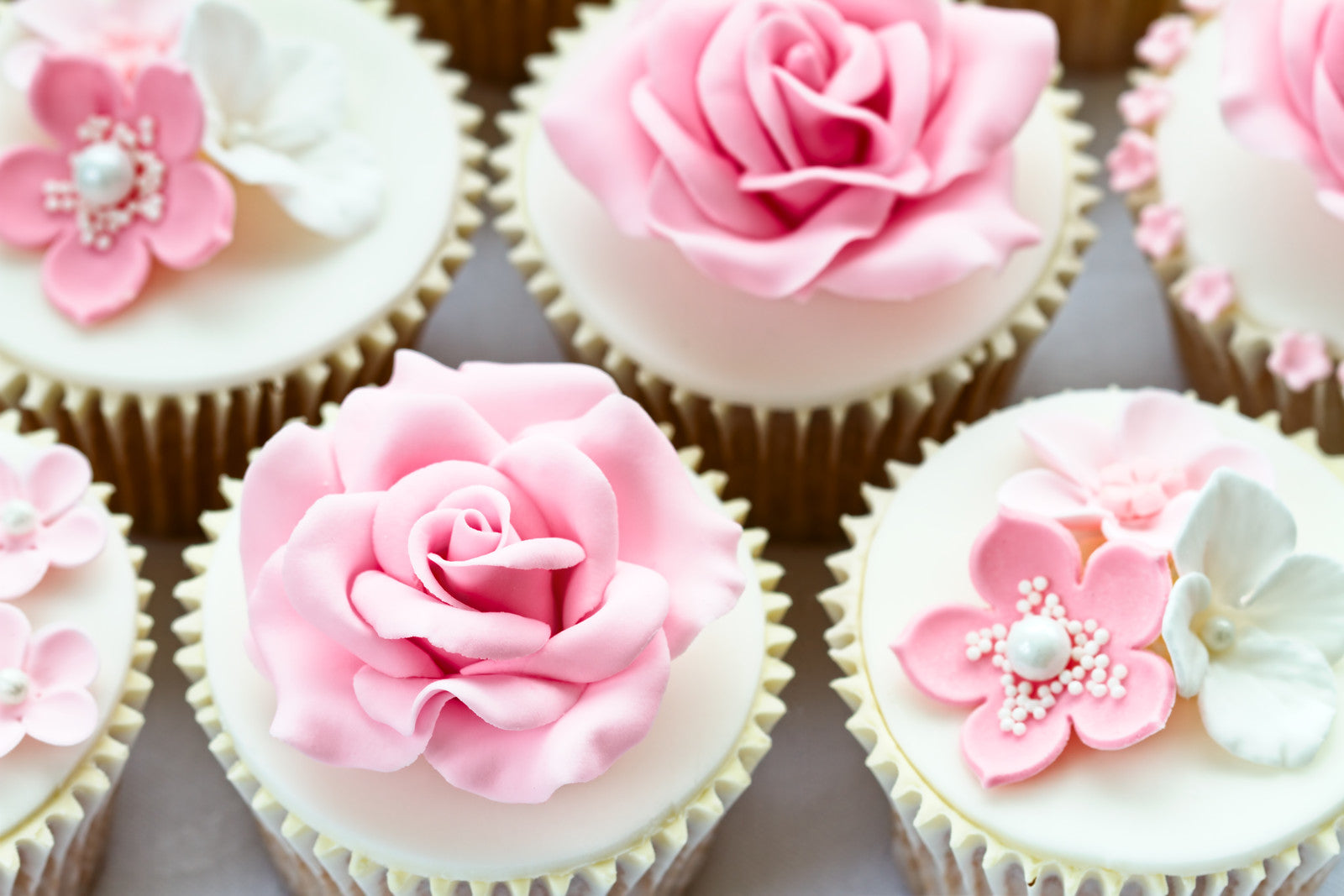 Cupcakes adorned with elegant pink and white fondant flowers, featuring roses and floral designs. The intricate decorations sit atop smooth white icing.