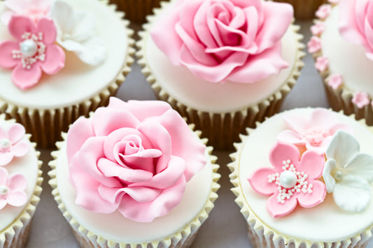 Cupcakes adorned with elegant pink and white fondant flowers, featuring roses and floral designs. The intricate decorations sit atop smooth white icing.